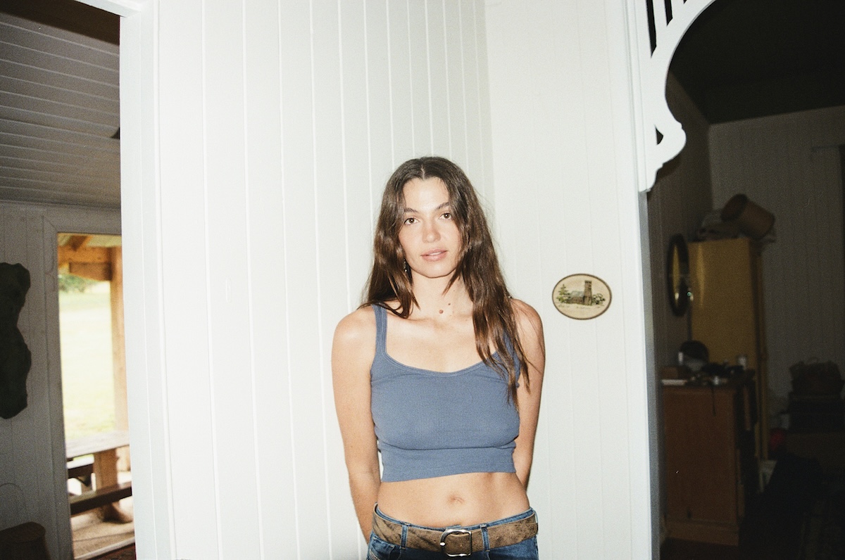 Australian singer-songwriter Kita Alexander wearing a blue tank top and jeans, standing against a white paneled wall inside a home.